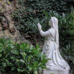 Figurka klęczącej kobiety z różańcami na rękach w skalnym ogrodzie